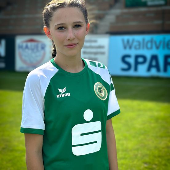 Eine Frau in einem grünen und weißen Fußballtrikot mit einem Logo auf der Brust steht auf einem grünen Feld. Sie schaut mit einem Lächeln nach vorne. Hinter ihr befinden sich verschwommene Werbungen für Waldvie und Spar.