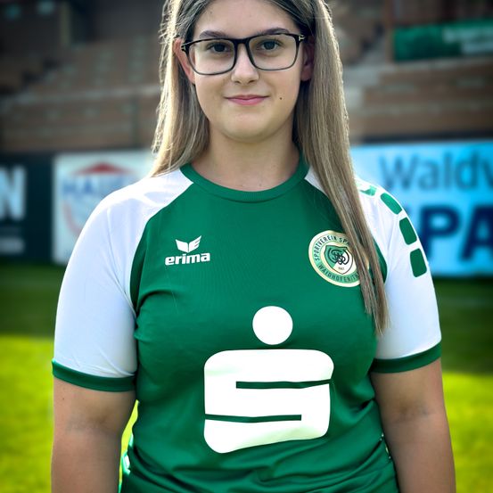 Eine Frau in einem grünen und weißen Trikot mit der Nummer 9 steht auf einem Feld, lächelt und schaut nach vorne. Dahinter ist ein verschwommener Blick auf ein Stadion mit einem grünen Banner.