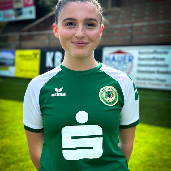 Eine Frau in einem grünen und weißen Fußballtrikot mit Logo und dem Wort Erima steht auf einem Feld.