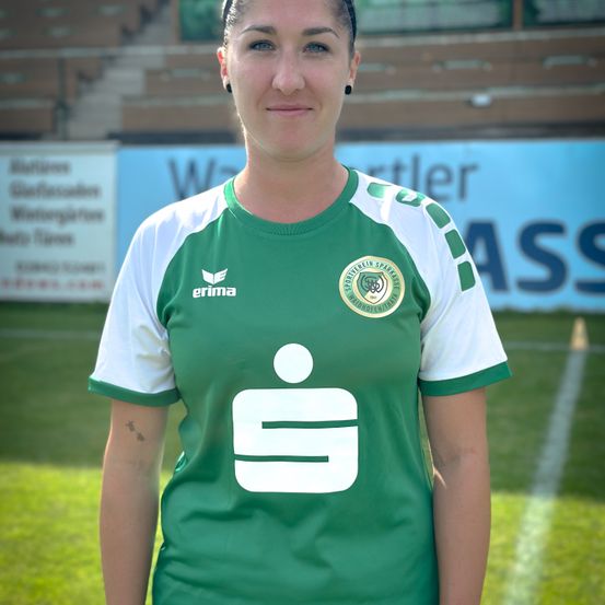 Eine Frau steht auf einem Fußballfeld und trägt ein grün-weißes Trikot mit Logo und Text. Hinter ihr ist ein verschwommener Hintergrund mit einem Zaun und Text auf Bannern.