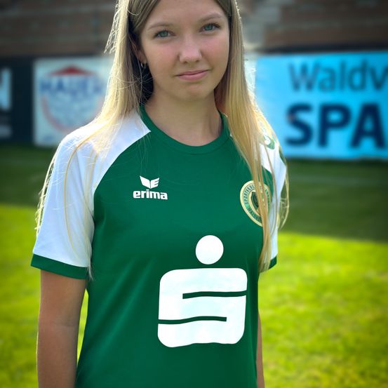 Eine Frau mit blonden Haaren trägt ein grünes und weißes Fußballtrikot und steht auf einem grünen Feld. Hinter ihr ist ein unscharfer Hintergrund mit einem Gebäude und einem Schild, auf dem Waldv SPA steht.