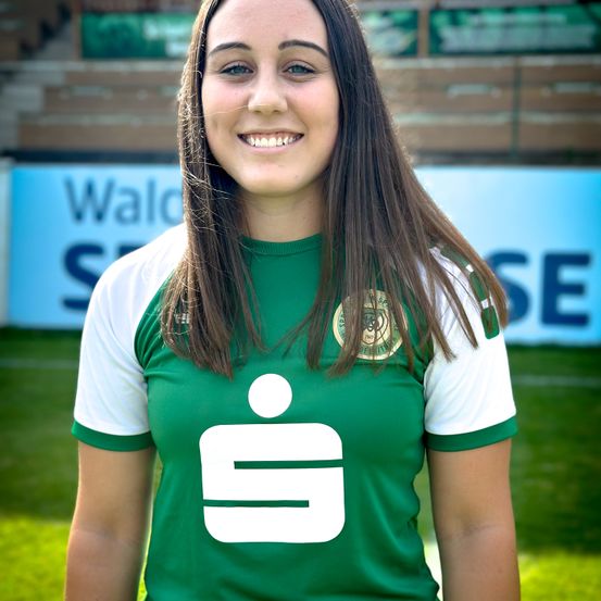 Eine Frau steht auf einem Feld und lächelt für ein Foto. Sie trägt ein grün-weißes Trikot mit einem Logo. Hinter ihr steht ein verschwommener Schild, auf dem 'Wald' und 'SE' steht.