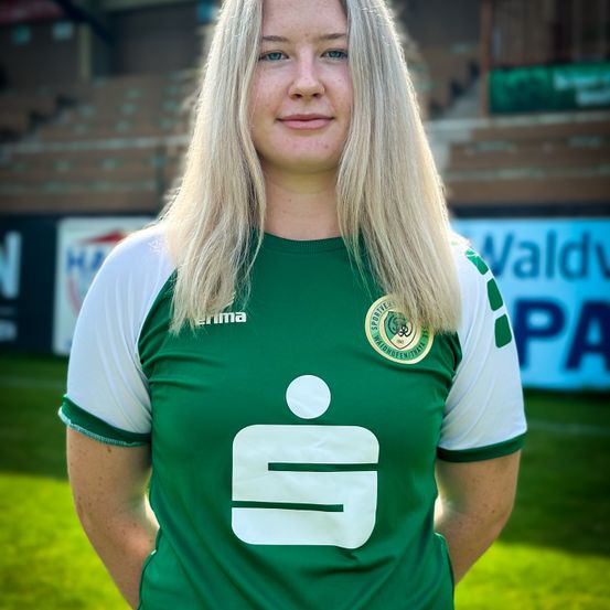 Eine Frau mit blonden Haaren steht auf einem grünen Feld und trägt ein grünes und weißes Fußballtrikot mit der Nummer 9. Sie schaut nach rechts.