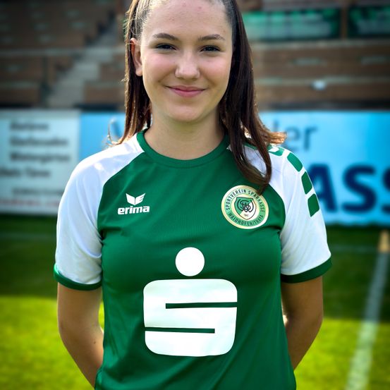Eine Frau steht auf einem Fußballfeld und trägt ein grün-weißes Trikot mit der Nummer 9 und einem Logo auf der linken Brust. Sie lächelt für ein Foto.