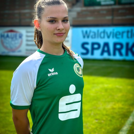 Eine Frau in einem grünen und weißen Fußballtrikot mit der Nummer 9 steht mit den Händen auf den Hüften auf einem Rasenfeld. Hinter ihr befindet sich ein Banner mit den Worten Waldviertler SPARK.
