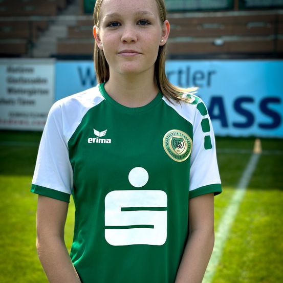 Eine Frau in einem grünen und weißen Fußballtrikot mit der Nummer '1' steht auf einem Feld. Das Trikot hat das Logo von 'SpVgg Reinfelden'. Der Hintergrund zeigt ein verschwommenes Banner mit Text.