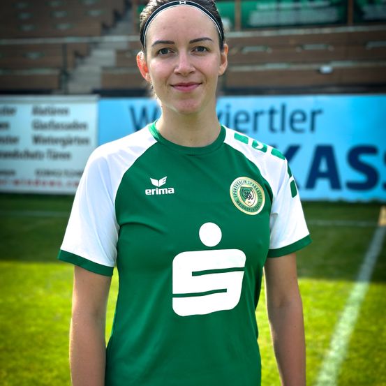 Eine Frau steht auf einem Fußballfeld, trägt ein grün-weißes Trikot mit Logo auf der Brust und einem Stirnband. Der Rasen ist grün und hat weiße Markierungen. Im Hintergrund sind unscharfe Werbungen zu sehen.