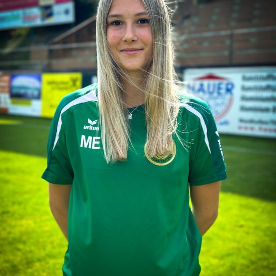 Eine Frau mit blonden Haaren steht auf einem Fußballfeld und trägt ein grünes Team-Trikot mit weißen Akzenten und einem Logo. Sie lächelt in die Kamera.