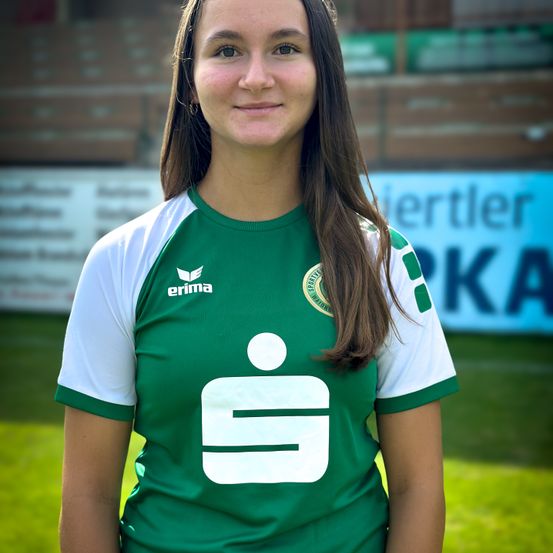 Eine Frau steht auf einem Sportplatz und trägt ein grün-weißes Trikot mit der Nummer 9. Sie lächelt und posiert für ein Foto. Hinter ihr ist ein verschwommener Blick auf einen Sportplatz mit einem blauen Banner.