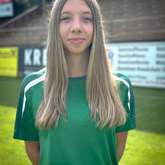 Eine junge Frau mit langen blonden Haaren steht auf einem grünen Feld und trägt ein grünes Shirt mit weißen Streifen. Hinter ihr ist eine Ziegelwand mit einem gelben Banner.