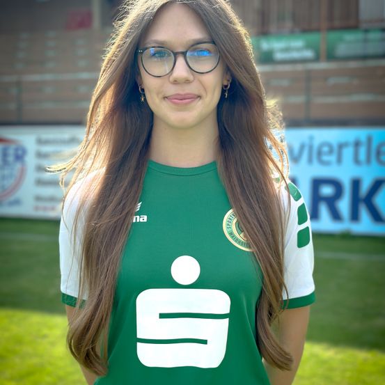 Eine Frau mit langen braunen Haaren, die ein grünes und weißes Sporttrikot mit Logo und Text trägt, steht auf einem Rasenfeld. Hinter ihr ist ein unscharfer Hintergrund mit Werbung.