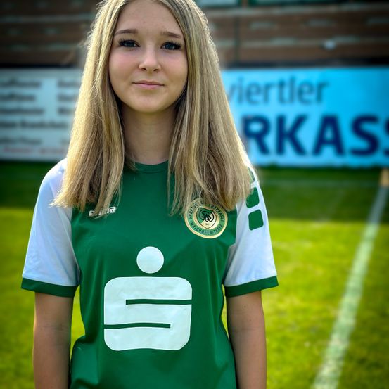 Eine Frau mit blonden Haaren steht auf einem Fußballfeld und trägt ein grünes und weißes Trikot mit einem Logo. Sie schaut mit einem leichten Lächeln nach vorne. Der Hintergrund umfasst einen Zaun und eine blaue Banner mit Text.