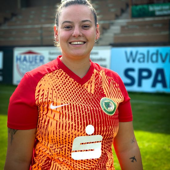 Eine Fußballspielerin mit einer Tätowierung am Arm steht lächelnd auf einem Fußballfeld. Sie trägt ein rotes und orangefarbenes Trikot mit einem Nike-Logo. Hinter ihr ist ein leeres Stadion mit einem Waldv SPA-Schild zu sehen.