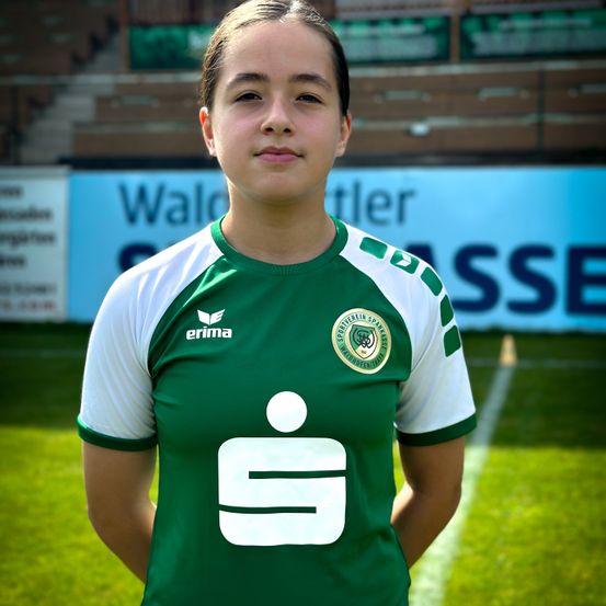 Eine Fußballspielerin in einem grün-weißen Trikot steht auf einem Feld, mit einem unscharfen Hintergrund, der eine Wand mit dem Wort 'Wald' und anderem Text zeigt.