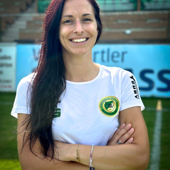 Eine Frau mit langen Haaren lächelt und trägt ein weißes T-Shirt mit Logo und grünem Emblem. Sie steht mit verschränkten Armen im Freien.