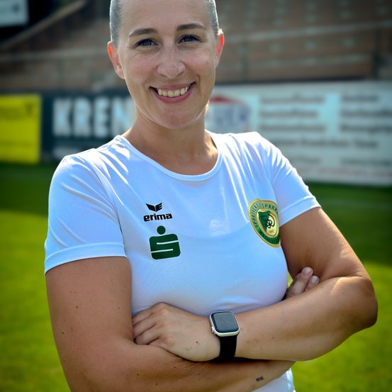 Eine Frau mit kurzen Haaren, die ein weißes Shirt mit dem Logo von Erima und einem grünen Aufnäher trägt, steht mit verschränkten Armen auf einem Rasenfeld.