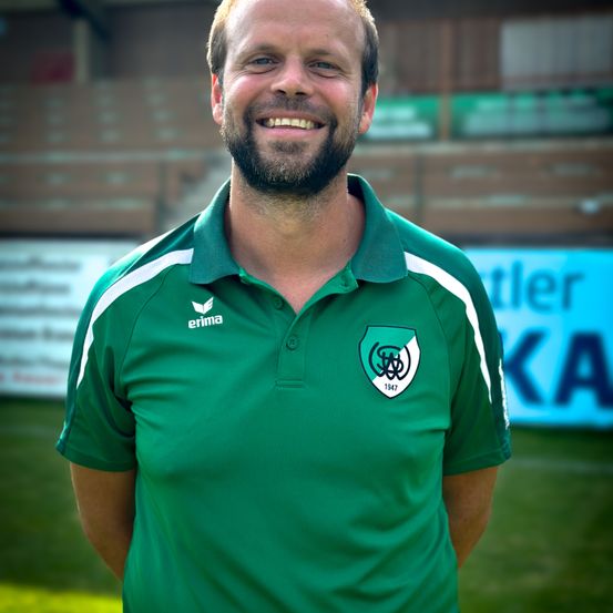 Ein Mann steht auf einem Sportplatz und lächelt. Er trägt ein grünes Polohemd mit weißem Kragen und weißen Streifen an den Ärmeln. Das Shirt hat ein Logo auf der linken Brust. Hinter ihm ist ein verschwommener Hintergrund eines Stadions mit leeren Sitzen.