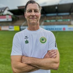 Ein Mann steht mit verschränkten Armen auf einem Fußballfeld, er trägt ein weißes T-Shirt mit dem Logo eines Sportvereins. Dahinter befindet sich ein verschwommenes Stadion mit leeren Sitzen.