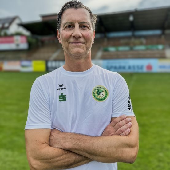 Ein Mann steht mit verschränkten Armen auf einem Fußballfeld, er trägt ein weißes T-Shirt mit dem Logo eines Sportvereins. Dahinter befindet sich ein verschwommenes Stadion mit leeren Sitzen.