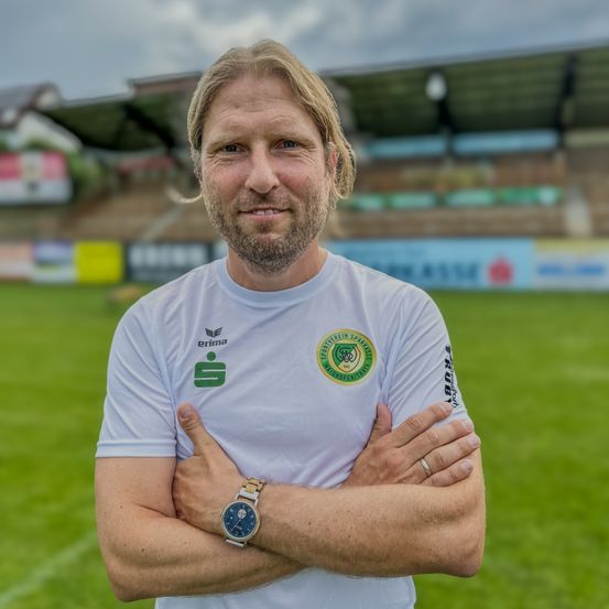 Ein Mann mit Bart steht mit verschränkten Armen auf einem grünen Feld, trägt ein weißes Shirt mit Logo und eine Uhr. Dahinter ist ein verschwommener Blick auf ein Stadion zu sehen.
