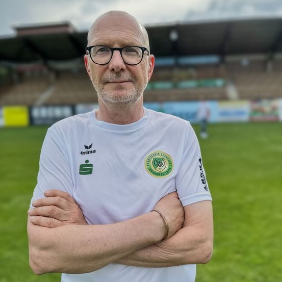 Ein älterer Mann steht mit verschränkten Armen in einem Fußballstadion. Er trägt ein weißes T-Shirt mit einem grünen Logo auf der linken Seite. Das Stadion hat leere Tribünen.