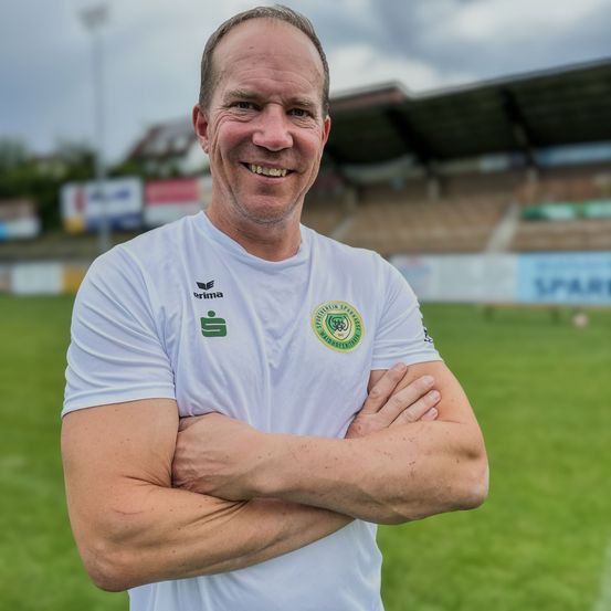 Ein lächelnder Mann mit verschränkten Armen posiert für ein Foto auf einem grünen Feld, trägt ein weißes Shirt mit Logos. Hinter ihm ein leeres Stadion mit Tribünen und einem bewölkten Himmel.