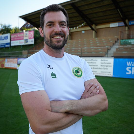 Ein Mann in einem weißen Shirt mit Logo steht auf einem Fußballfeld mit verschränkten Armen. Dahinter sind leere Tribünen.