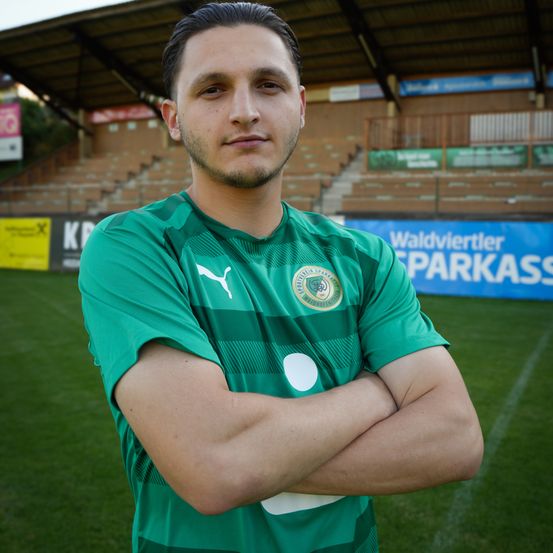 Ein Fußballspieler steht auf einem Feld und trägt ein grünes Trikot mit einem Logo auf der Brust. Das Feld hat ein blaues Banner, das Waldviertler Sparkasse steht.