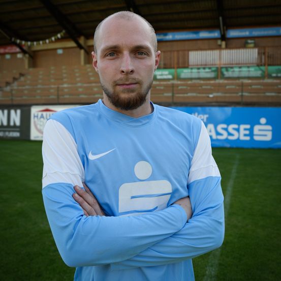 Ein Mann in einem blauen Nike-Fußballtrikot steht mit verschränkten Armen auf einem Feld, mit einem Banner hinter ihm, das 'ASSÉS' steht.