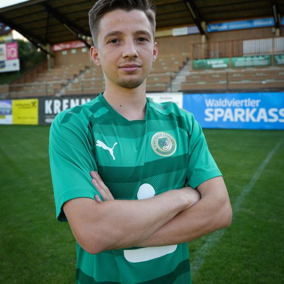 Ein Mann steht auf einem Fußballfeld mit verschränkten Armen und trägt ein grünes Trikot mit Logo auf der Brust. Das Spielfeld hat weiße Linien und hinter ihm befindet sich ein teilweise leeres Stadion.