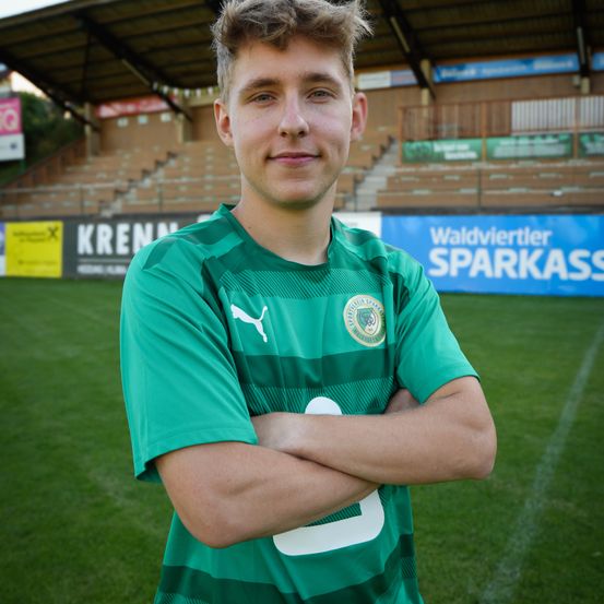 Ein lächelnder Fußballspieler steht auf einem Feld, trägt ein grünes Trikot mit Logo und hat die Arme verschränkt. Hinter ihm steht auf einem großen Banner Waldviertler Sparkasse.