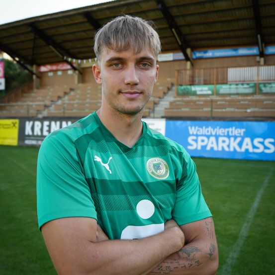 Ein Mann steht in einem Fußballstadion und trägt ein grünes Trikot mit weißen und grünen Streifen. Er lächelt für die Kamera. Hinter ihm befindet sich ein blauer Banner mit weißem Text.