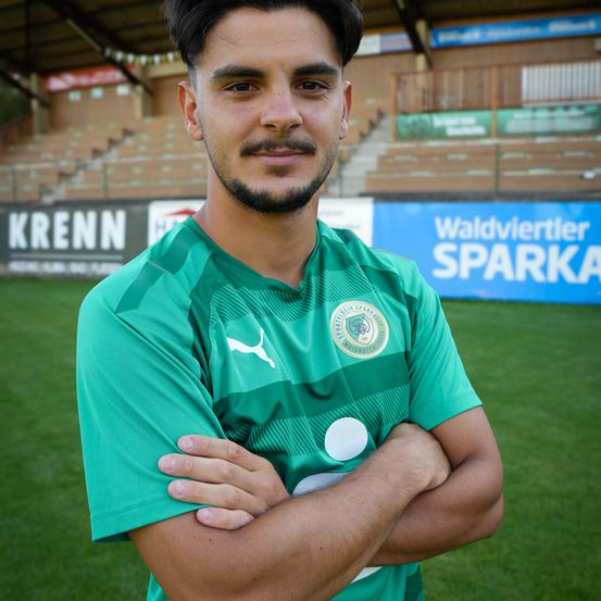 Ein Fußballspieler posiert vor einem Feld, trägt ein grünes Shirt mit Puma-Logo und einem weißen Kreis auf der Brust. Dahinter befindet sich ein Schild für Waldviertler Sparka.
