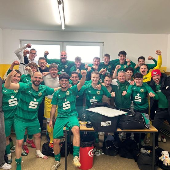 Ein Fußballteam in grünen Trikots feiert einen Sieg in der Umkleidekabine, hebt die Fäuste und lächelt, mit Taschen und einem Tisch im Hintergrund.