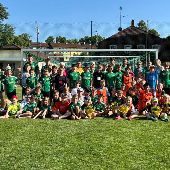 Eine Gruppe von Kindern und Erwachsenen in grünen und orangefarbenen Trikots posiert für ein Foto auf einem Fußballfeld. Das Fußballtor ist im Hintergrund zu sehen.