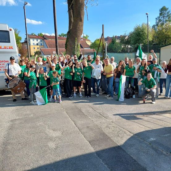 Eine Gruppe von Menschen in grünen Trikots posiert für ein Foto, einige mit Flaggen, in der Nähe eines Busses und Bäumen.