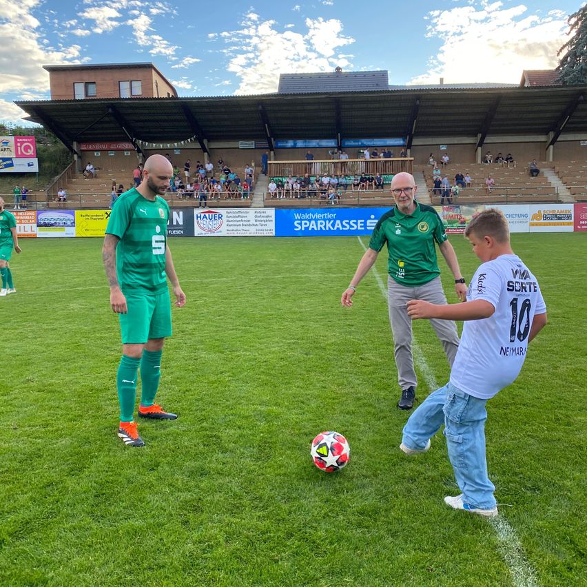 Auf einem Fußballplatz tritt ein Junge einen Ball, während zwei Männer zusehen. Ein Mann trägt ein grünes Trikot, der andere ein weißes Hemd. Im Hintergrund befinden sich Tribünen mit Zuschauern.
