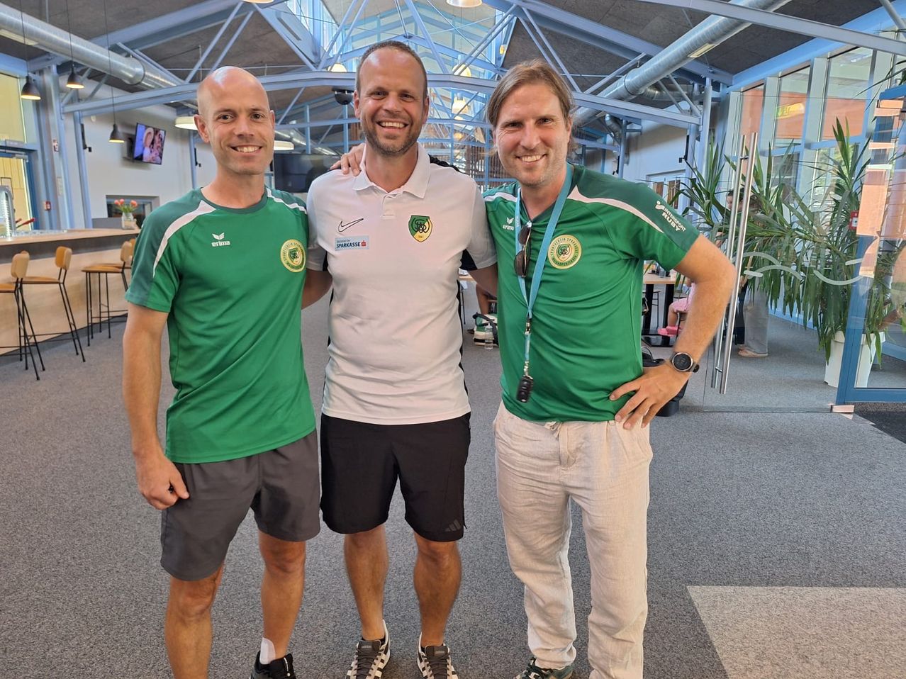Drei Männer in Sportbekleidung, zwei in Grün und einer in Weiß, stehen lächelnd in einer Lobby. Sie sind wahrscheinlich Athleten oder Trainer.