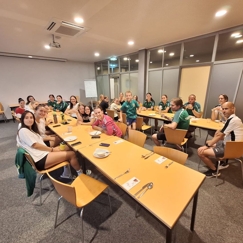 Eine Gruppe von Menschen in Sportuniformen sitzt an einem langen Tisch in einem Besprechungsraum. Teller, Besteck und Getränke stehen auf dem Tisch.