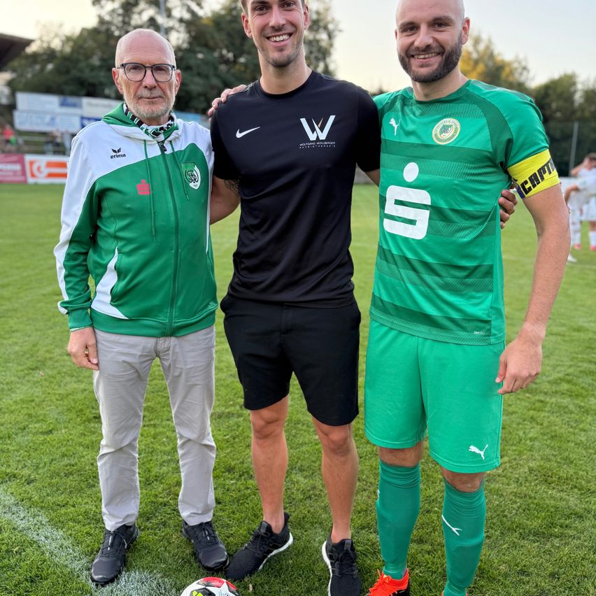 Drei Männer stehen auf einem Fußballfeld. Der Mann in der Mitte trägt ein schwarzes Nike-Trikot, während die anderen grüne Trikots tragen. Sie alle lächeln und scheinen für ein Foto zu posieren.