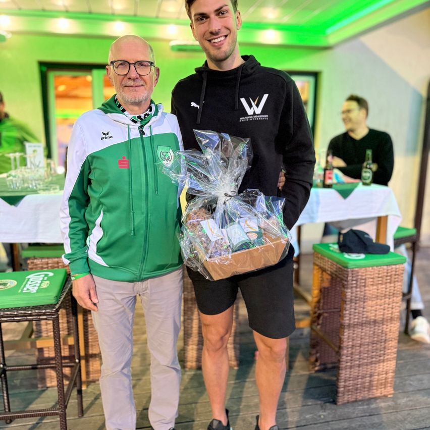 Zwei Männer stehen zusammen und halten einen Geschenkkorb. Einer trägt eine grüne Jacke, der andere einen schwarzen Kapuzenpullover. Sie sind in einem dekorierten Raum mit Tischen und Stühlen.