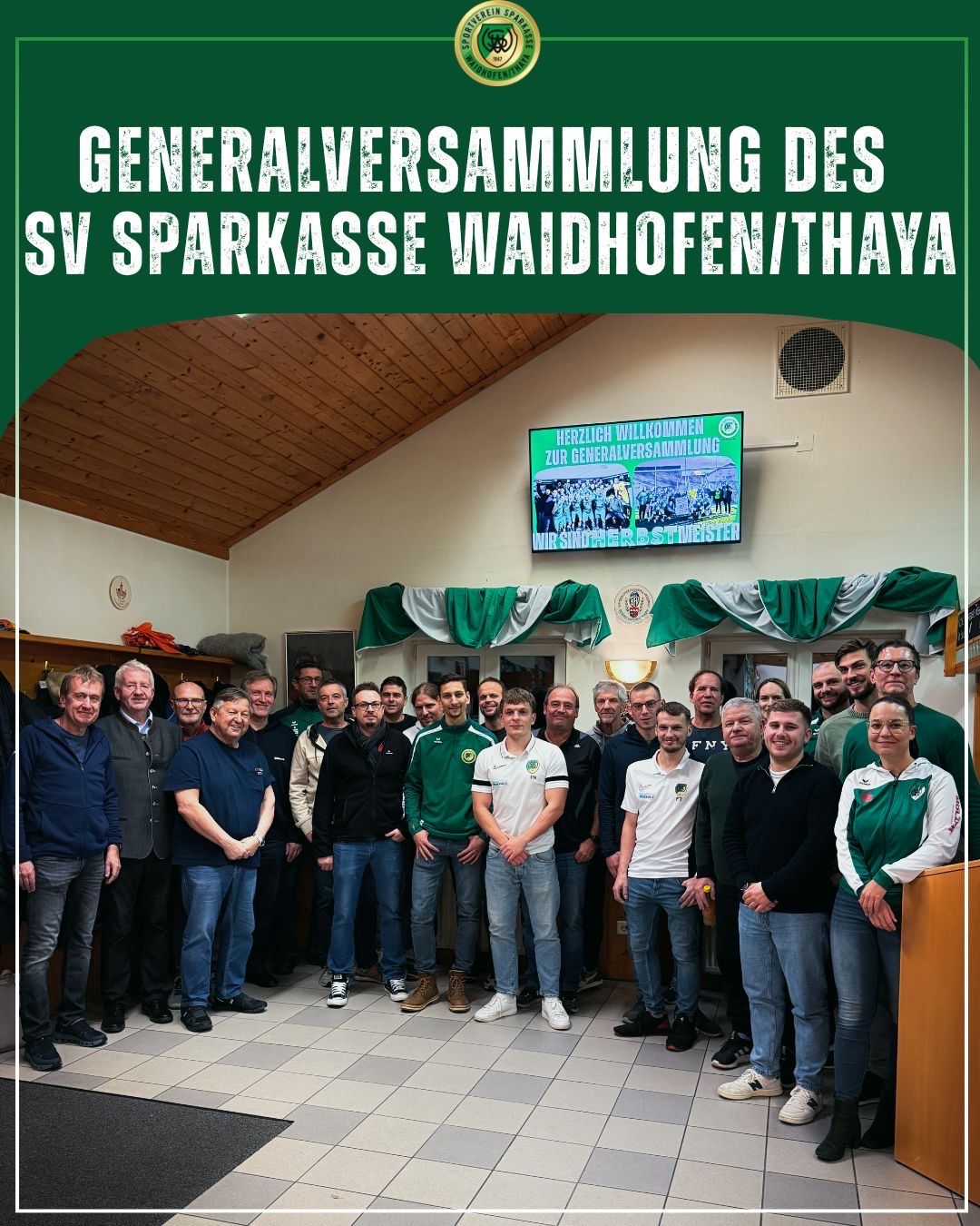 Eine Gruppe von Männern und Frauen in passenden grünen und weißen Trikots steht lächelnd für das Foto zusammen in einem Raum. Ein Monitor zeigt eine Nachricht an, die sie zur Generalversammlung von SV Sparkasse Waidhofen/Thaya begrüßt.