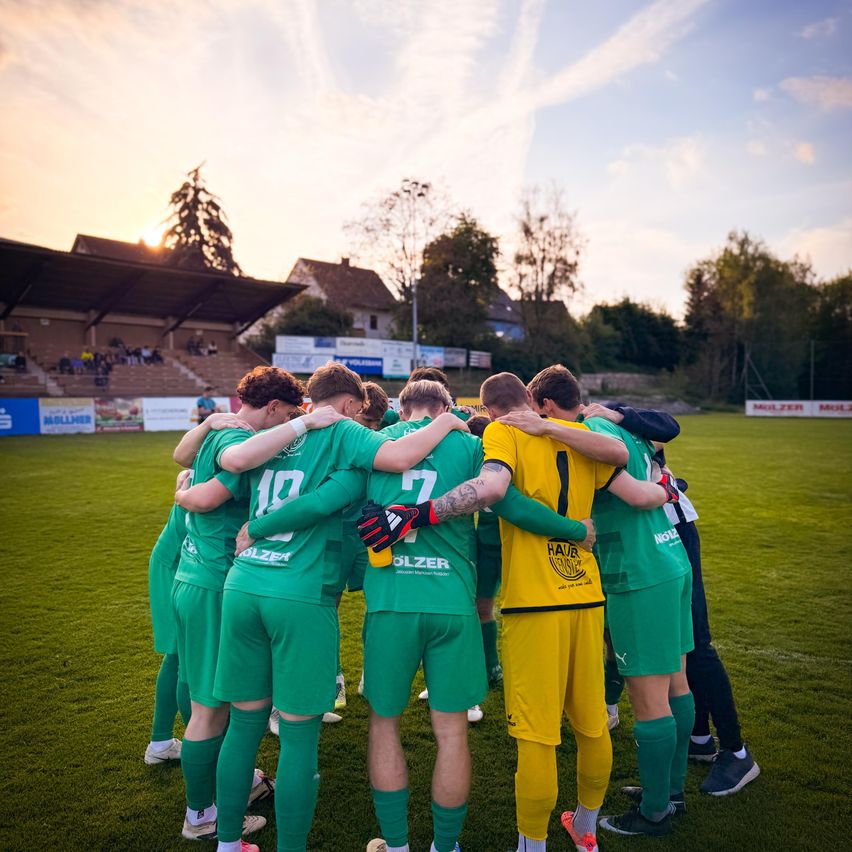 Fußballspieler in grünen Trikots, mit Armen umeinander, stehen auf einem Feld bei Sonnenuntergang, mit einem Publikum im Hintergrund.
