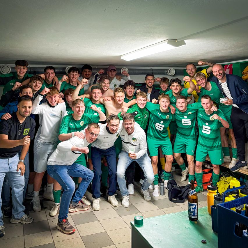 Eine Fußballmannschaft in grünen Trikots feiert in einer Umkleidekabine mit Bierflaschen und Snacks, unter einer Deckenleuchte.