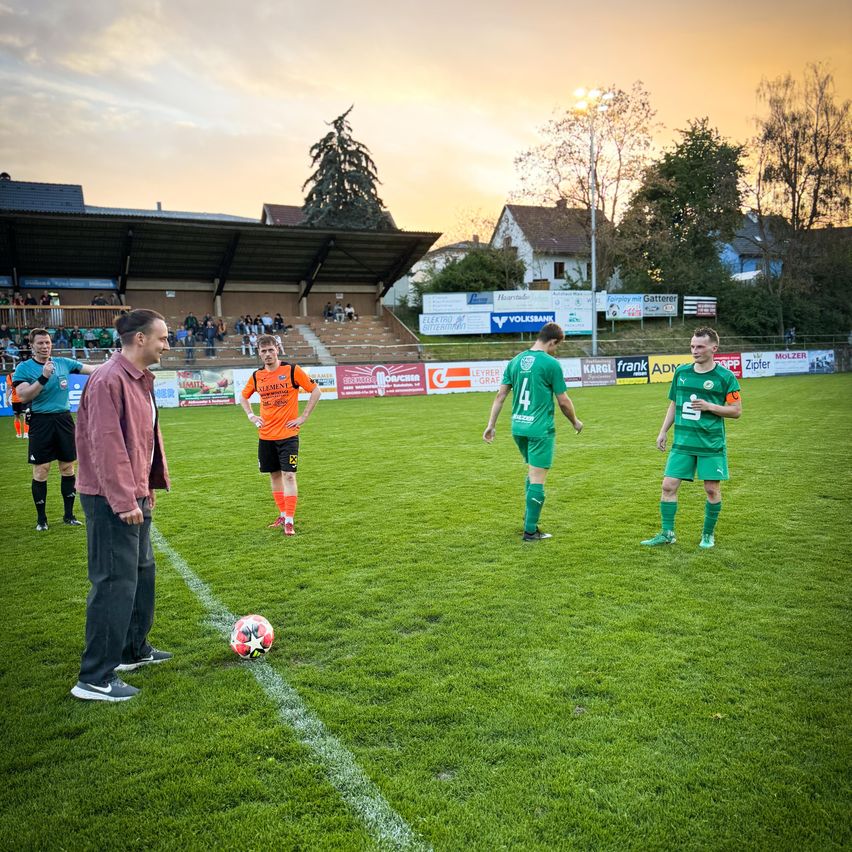 Fußballspieler in grünen und orangefarbenen Trikots unterhalten sich in der Nähe der Feldmitte, ein Schiedsrichter und ein Ball sind in der Nähe.