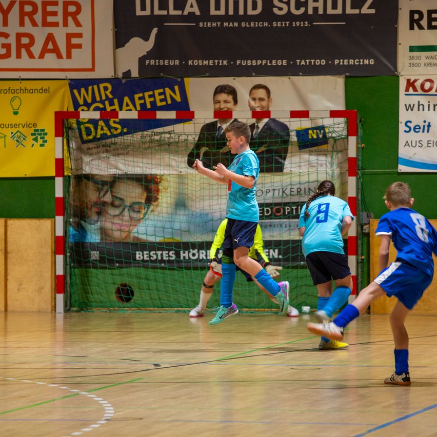 Junge Spieler in Trikots spielen Hallenfußball, ein Spieler springt, um den Ball zu köpfen. Der Platz ist von Plakaten und einem grünen Netz umgeben.