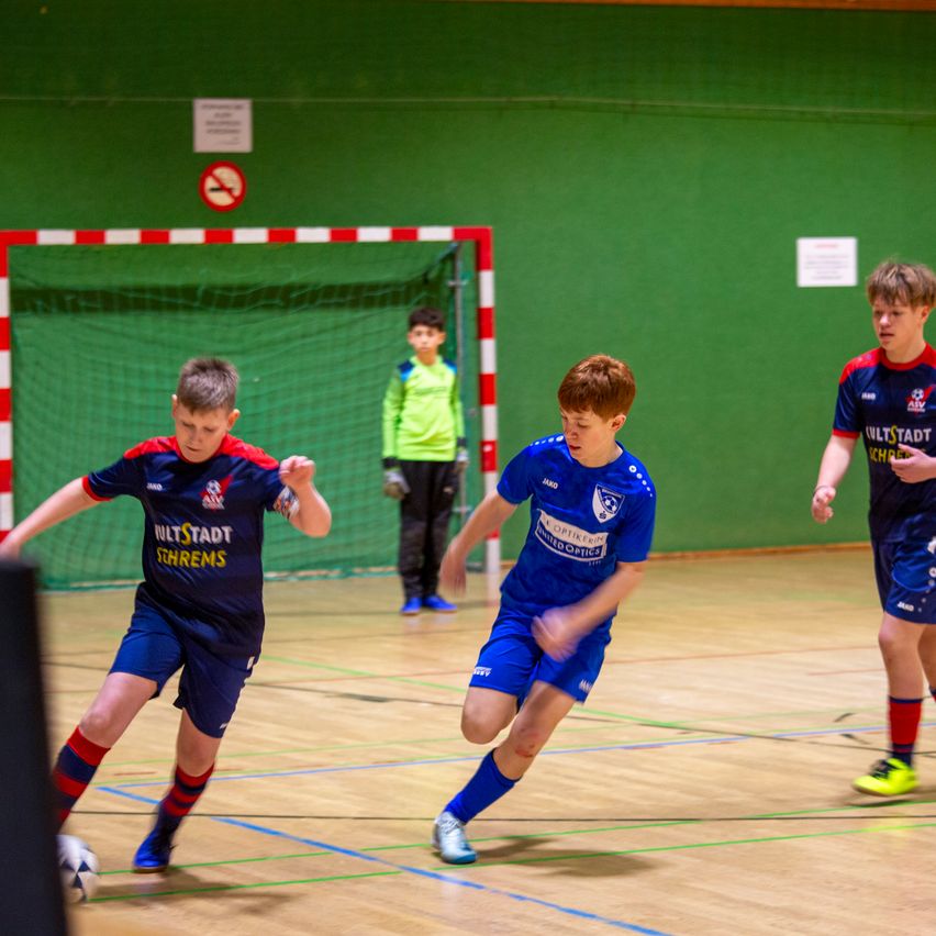 Vier Jungen spielen Hallenfußball. Zwei jagen einem Ball hinterher, während zwei andere in der Nähe des Tores stehen.