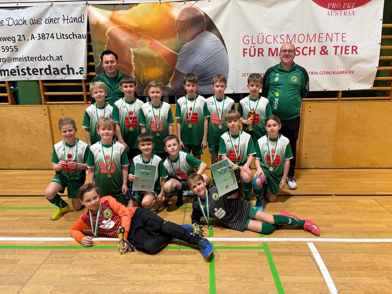 Eine Jugendsportmannschaft posiert auf einem Feld für ein Foto, trägt grüne und weiße Trikots, Medaillen und hält Urkunden. Zwei Erwachsene stehen hinter ihnen.