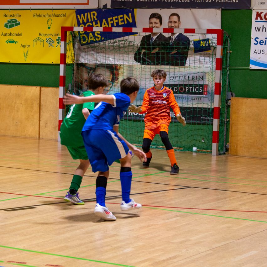 Drei Kinder spielen Hallenfußball, zwei in Blau und eines in Orange. Sie sind in der Nähe eines Tores mit einer Werbung an der Wand.
