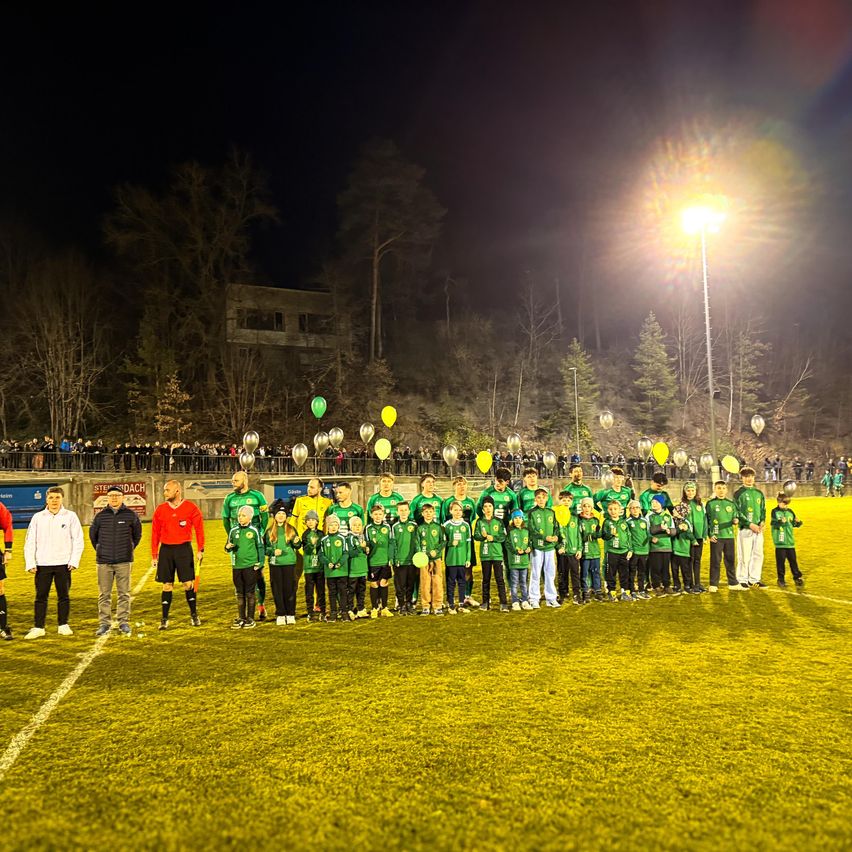 A soccer team in green uniforms poses for a photo on a field at night, with spectators and balloons in the background.